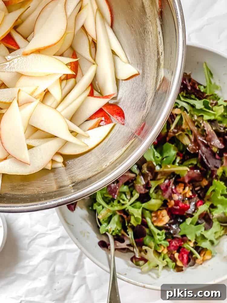 Harvest Pear and Gorgonzola Salad 6 A hand is adding sliced pears into a large bowl already containing mixed greens, cranberries, and nuts.
