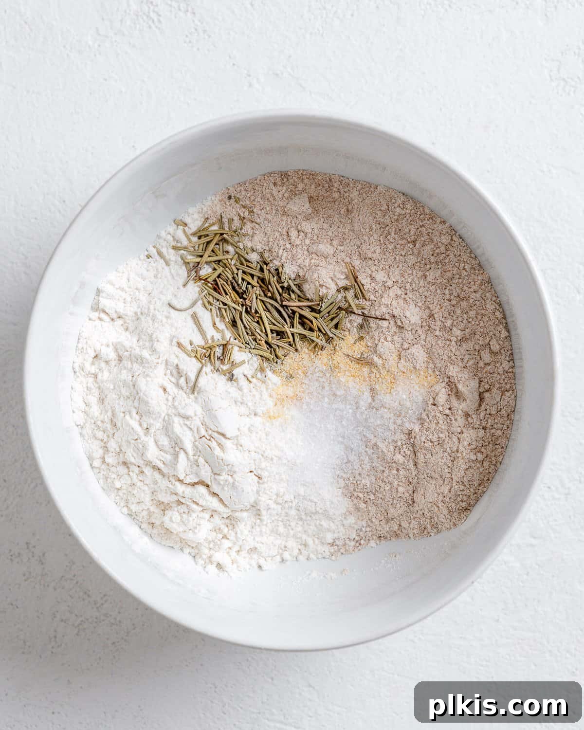 Rustic Rosemary Vegan Crisps 5 Process of adding flour and spices to a white bowl on a white background.