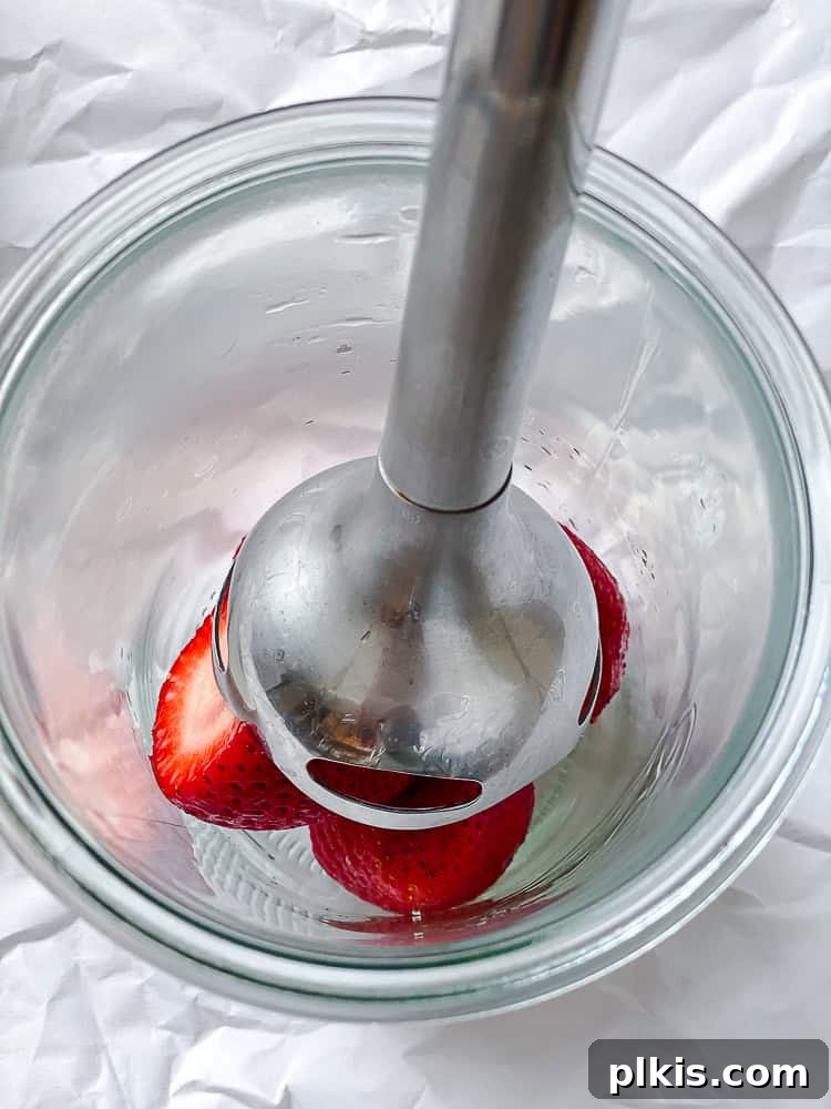 Effortless Strawberry Glaze for All Your Sweet Treats 4 A process shot showing an immersion blender pureeing fresh strawberries in a cup, creating a smooth mixture.
