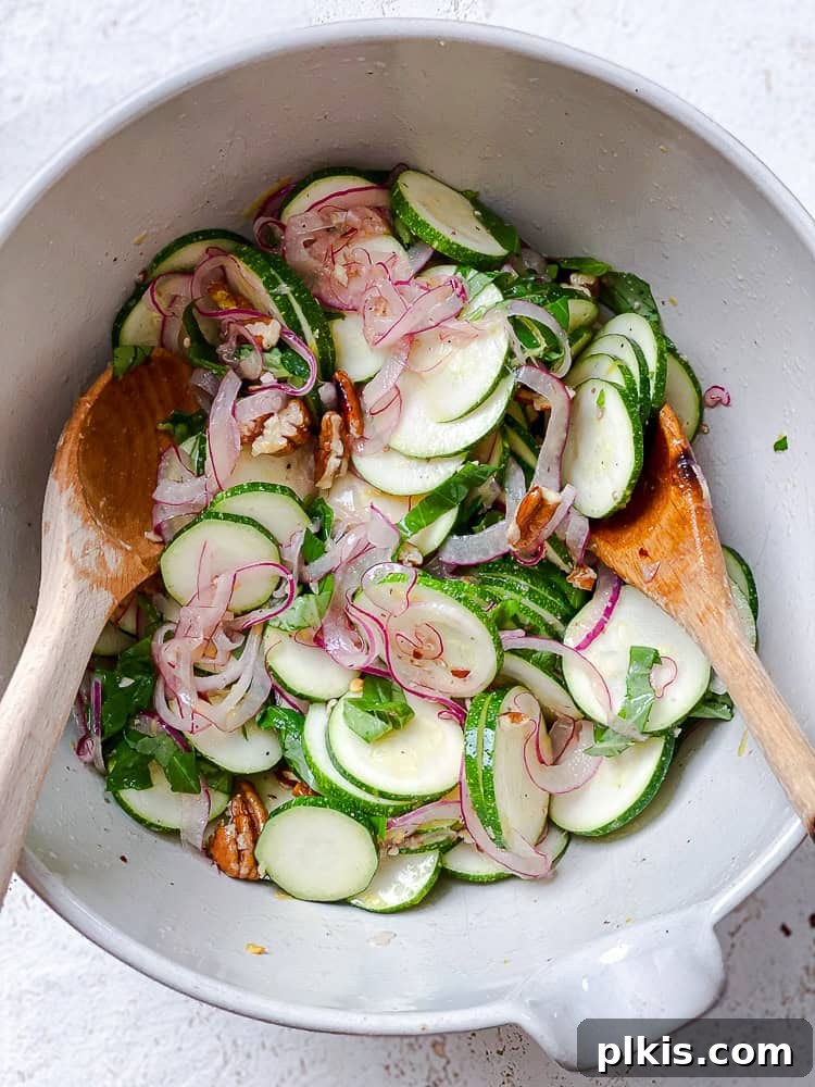 Zesty Zucchini Salad 9 Two wooden spoons are used for a final toss of all zucchini salad ingredients together in a large white bowl, ensuring even coating.
