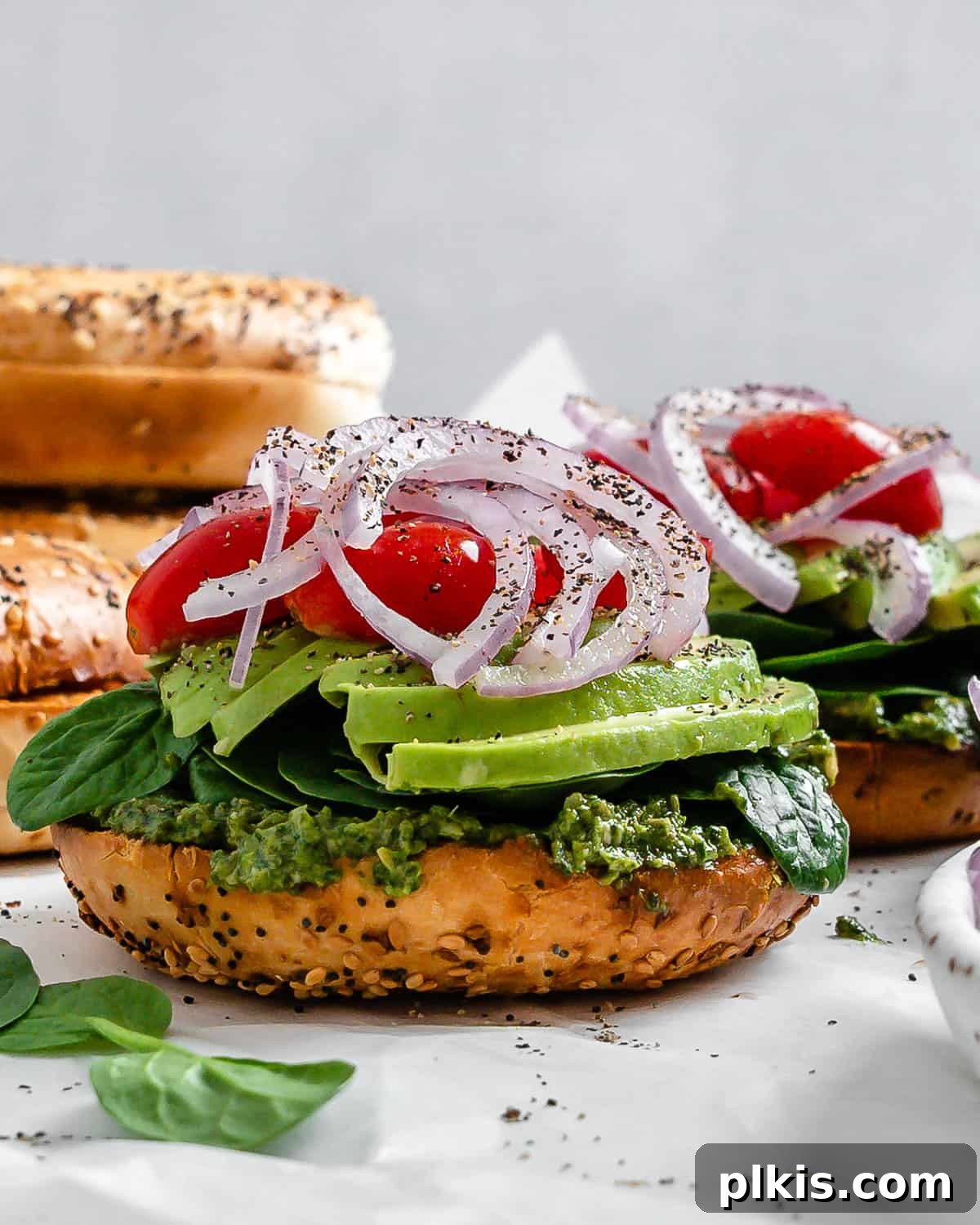 Two vibrant vegan pesto bagel sandwiches, piled high with fresh vegetables, on a clean white surface with a minimalist background.