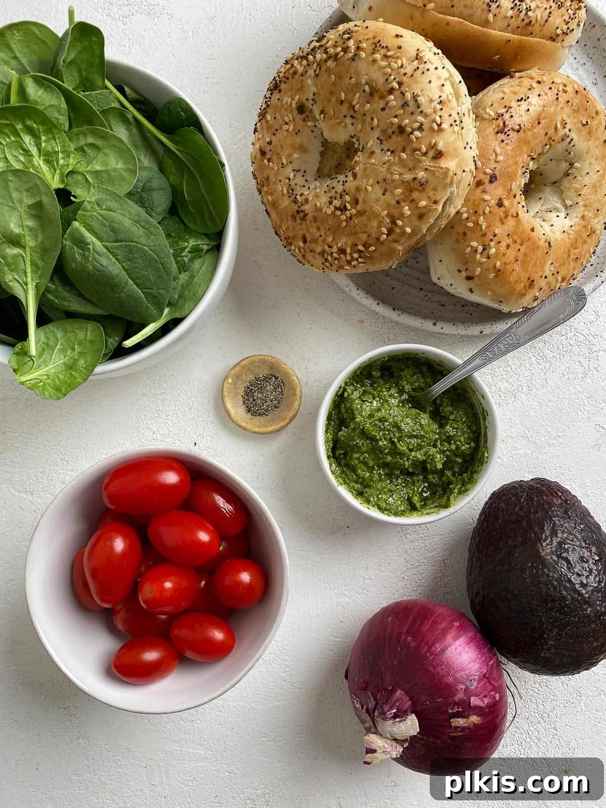 Fresh ingredients for a vegan pesto bagel sandwich laid out neatly on a white background, including bagels, pesto, spinach, avocado, tomatoes, and red onion.