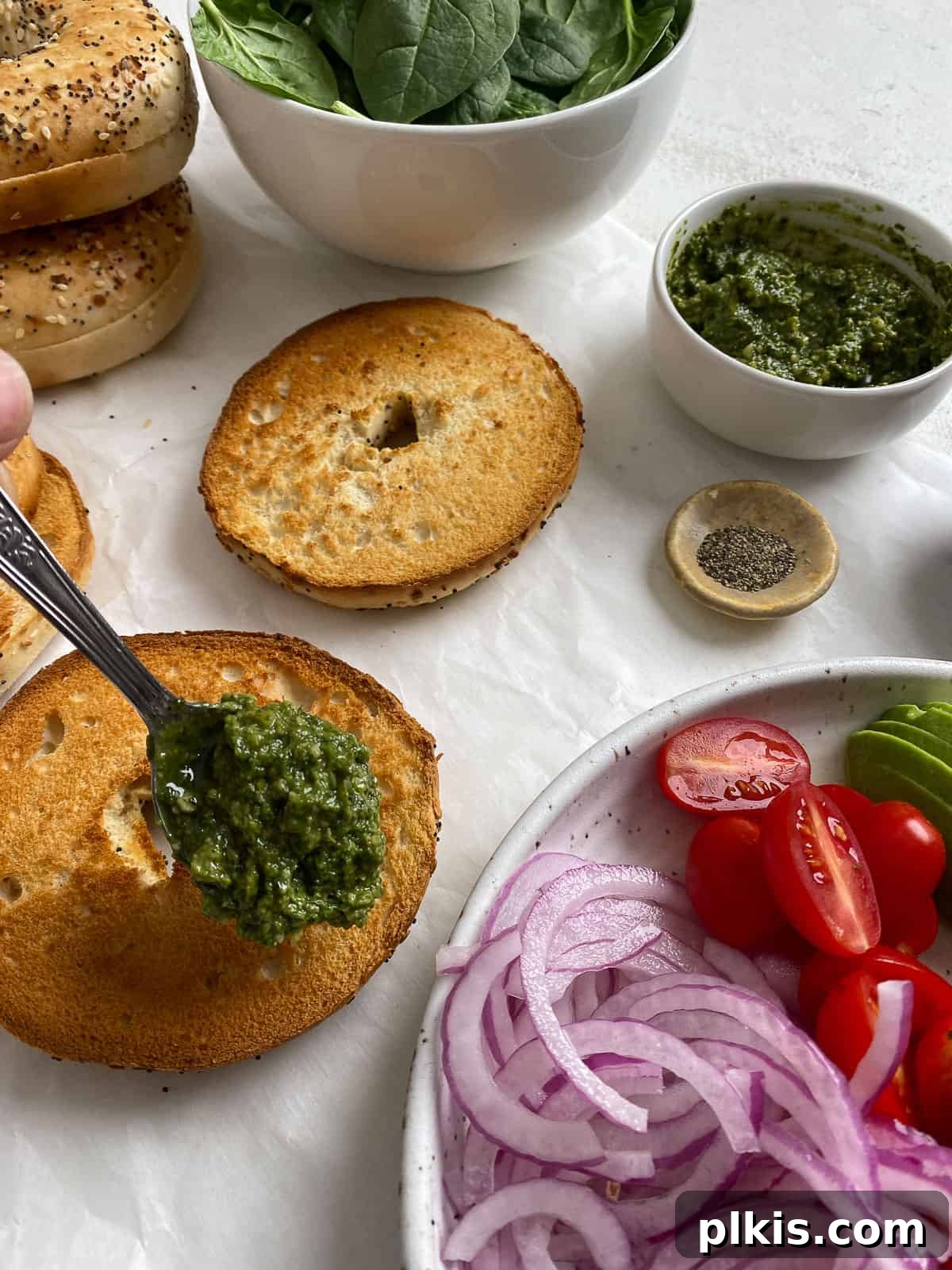 Process shot: A toasted bagel half is being spread with bright green pesto on a white surface, with fresh ingredients in the background.