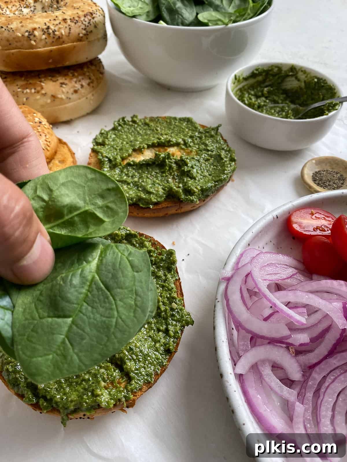 Process shot: Fresh baby spinach leaves are being layered onto a pesto-covered bagel half, with other fresh ingredients visible.