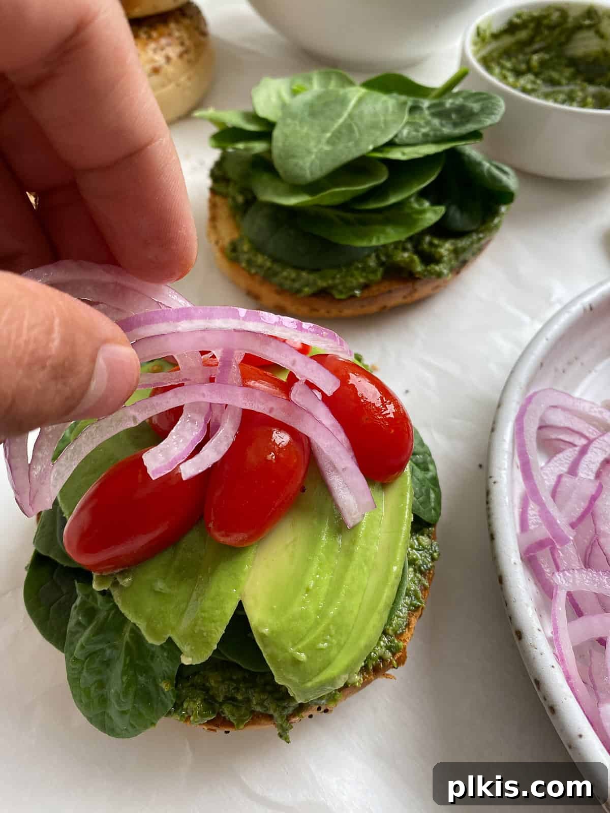 Process shot: Thinly sliced red onion is being placed on top of a vegan pesto bagel sandwich layered with spinach and avocado.