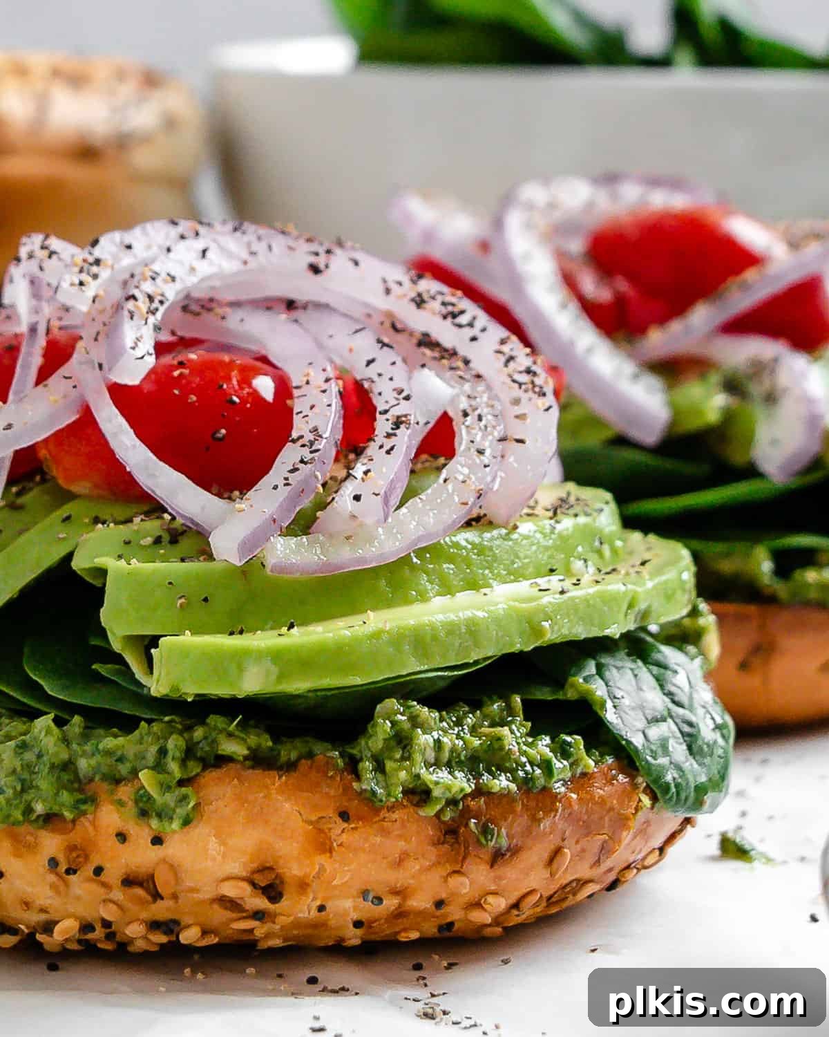 Two open-faced vegan pesto bagel sandwiches displayed, showcasing vibrant layers of green pesto, spinach, avocado, tomatoes, and red onion, on a bright white surface.