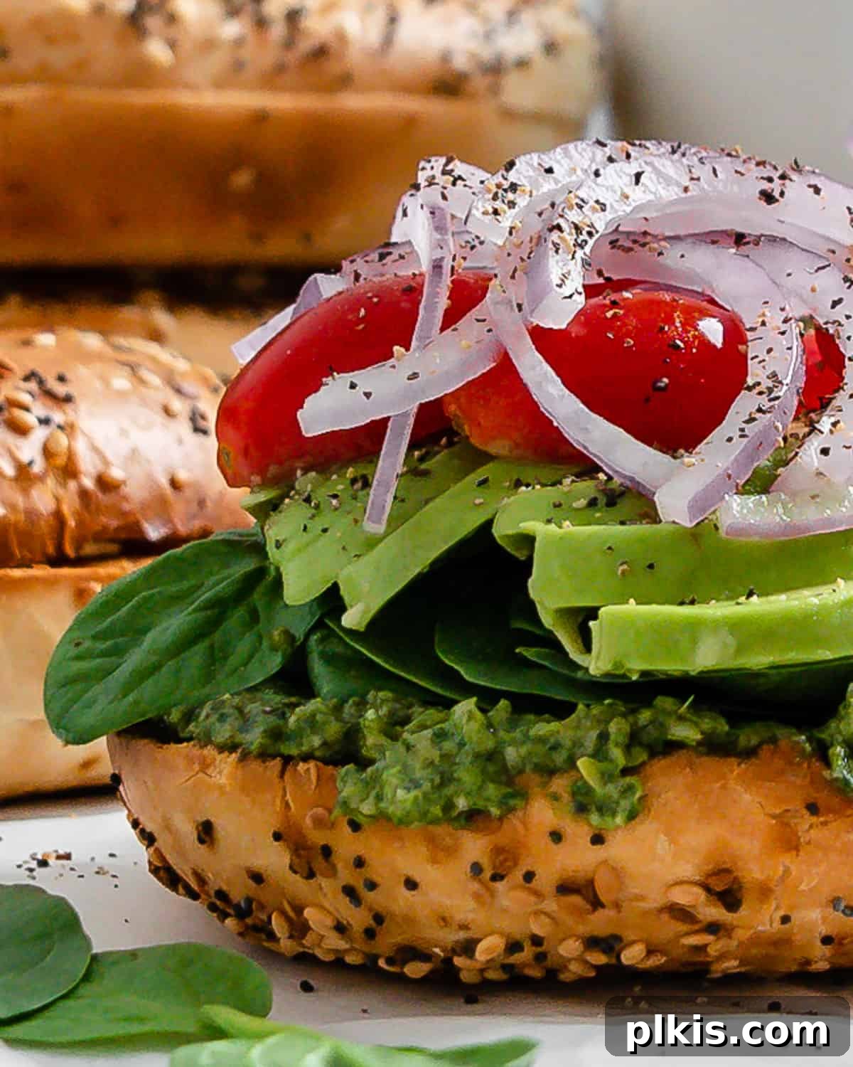 A close-up shot of a single vegan pesto bagel sandwich, showcasing the vibrant layers of ingredients on a toasted bagel half, presented on a white surface.