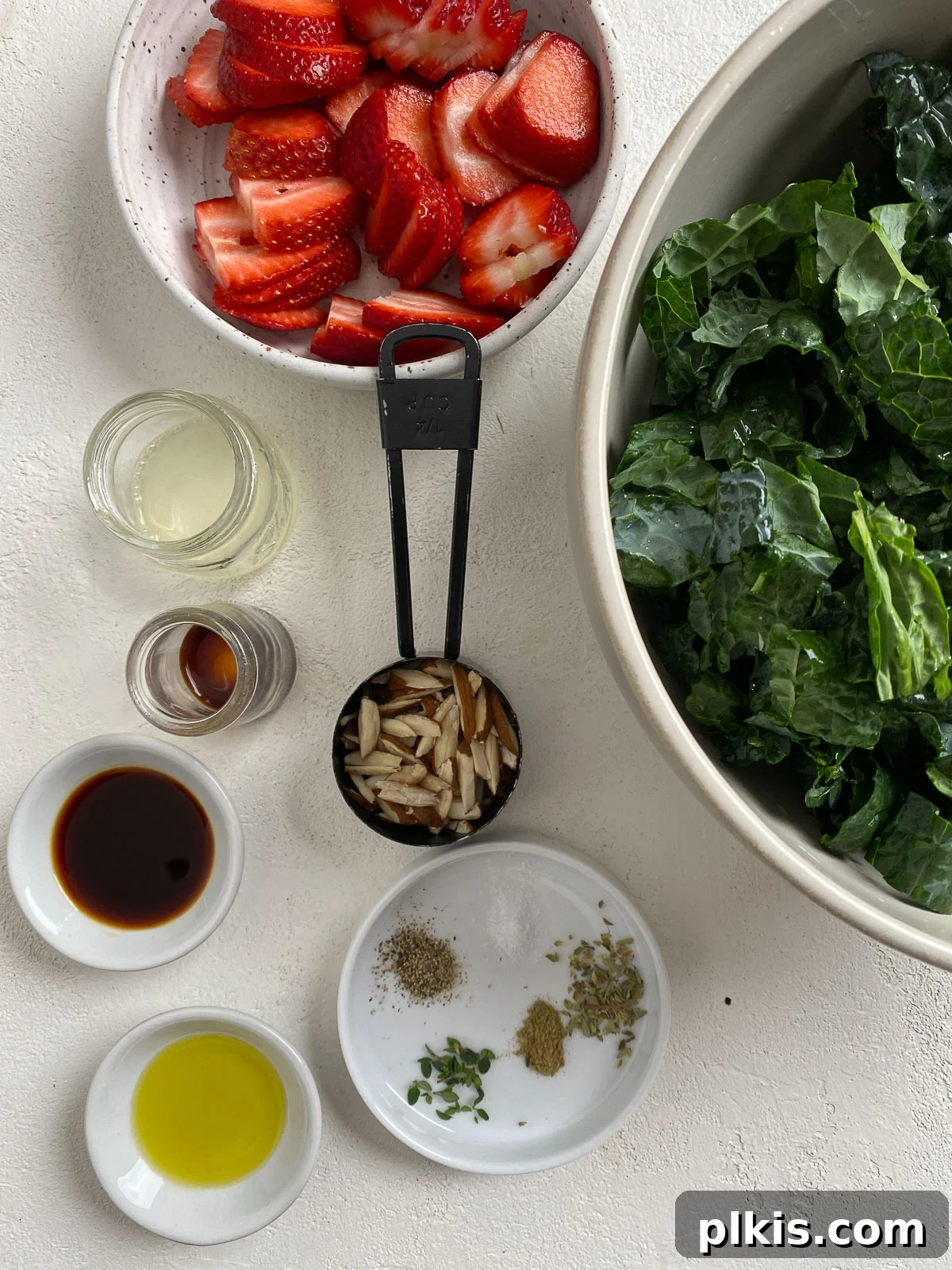 Berry Burst Kale Salad 3 ingredients for Strawberry Kale Salad measured out against a light surface, including fresh kale, sliced strawberries, almonds, olive oil, balsamic vinegar, Dijon mustard, and spices