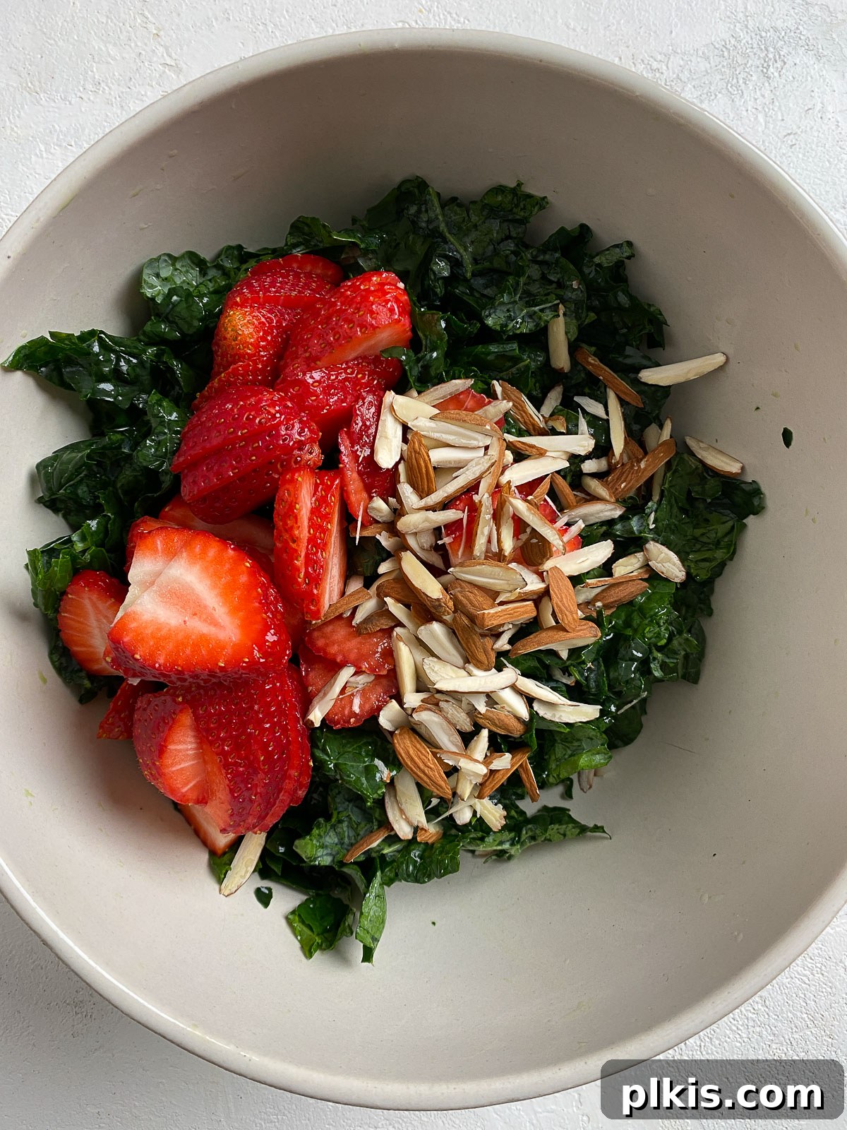 Berry Burst Kale Salad 4 process showing addition of chopped kale, sliced strawberries, and slivered almonds into a large white bowl for salad preparation