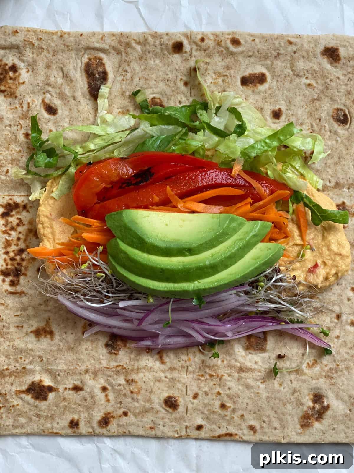 Effortless Hummus Roll Ups 5 Process shot illustrating various fresh vegetables and sliced avocado being neatly layered onto a hummus-spread tortilla, preparing it for rolling.