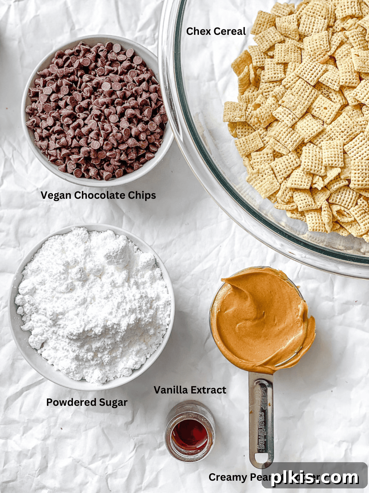 A selection of ingredients laid out on a white surface, including Chex cereal, a bag of vegan chocolate chips, a jar of creamy peanut butter, a bottle of vanilla extract, and a bowl of powdered sugar, all ready for making Easy Muddy Buddies [Puppy Chow].