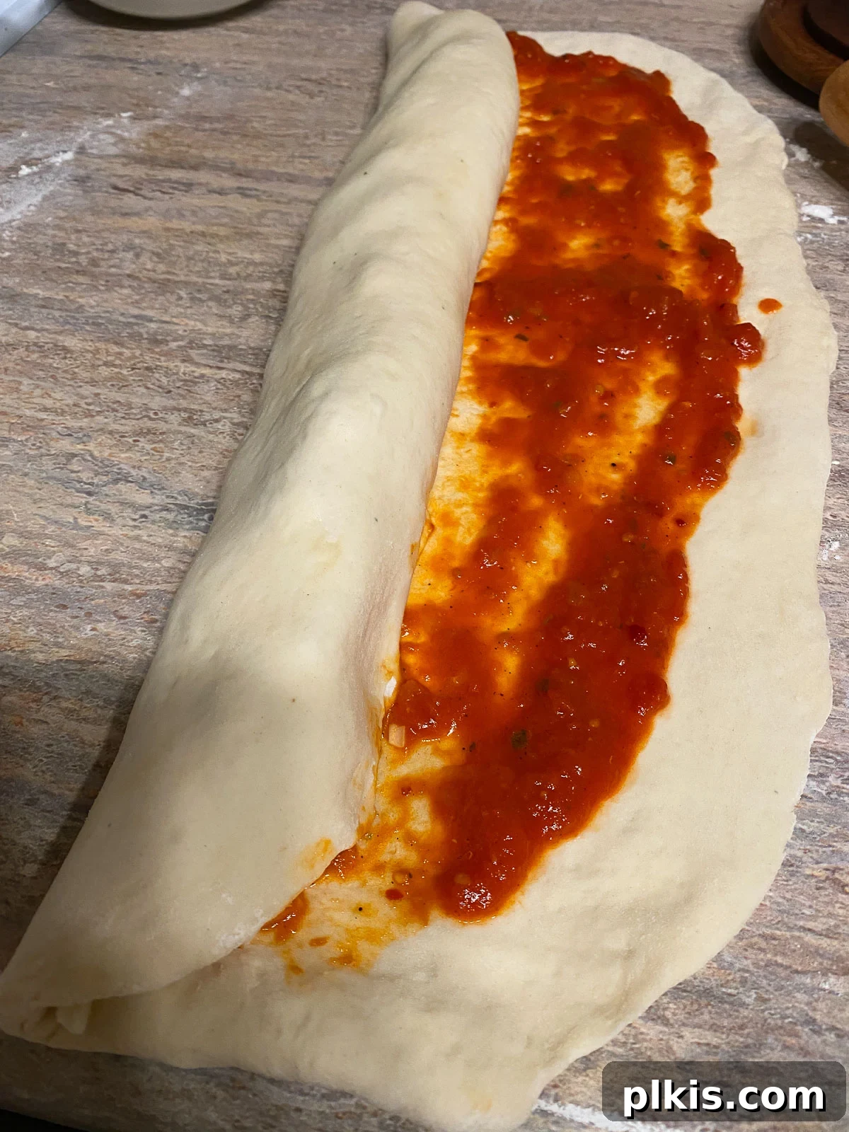 The pizza dough being carefully rolled up into a log from the bottom edge, creating a spiral of fillings.