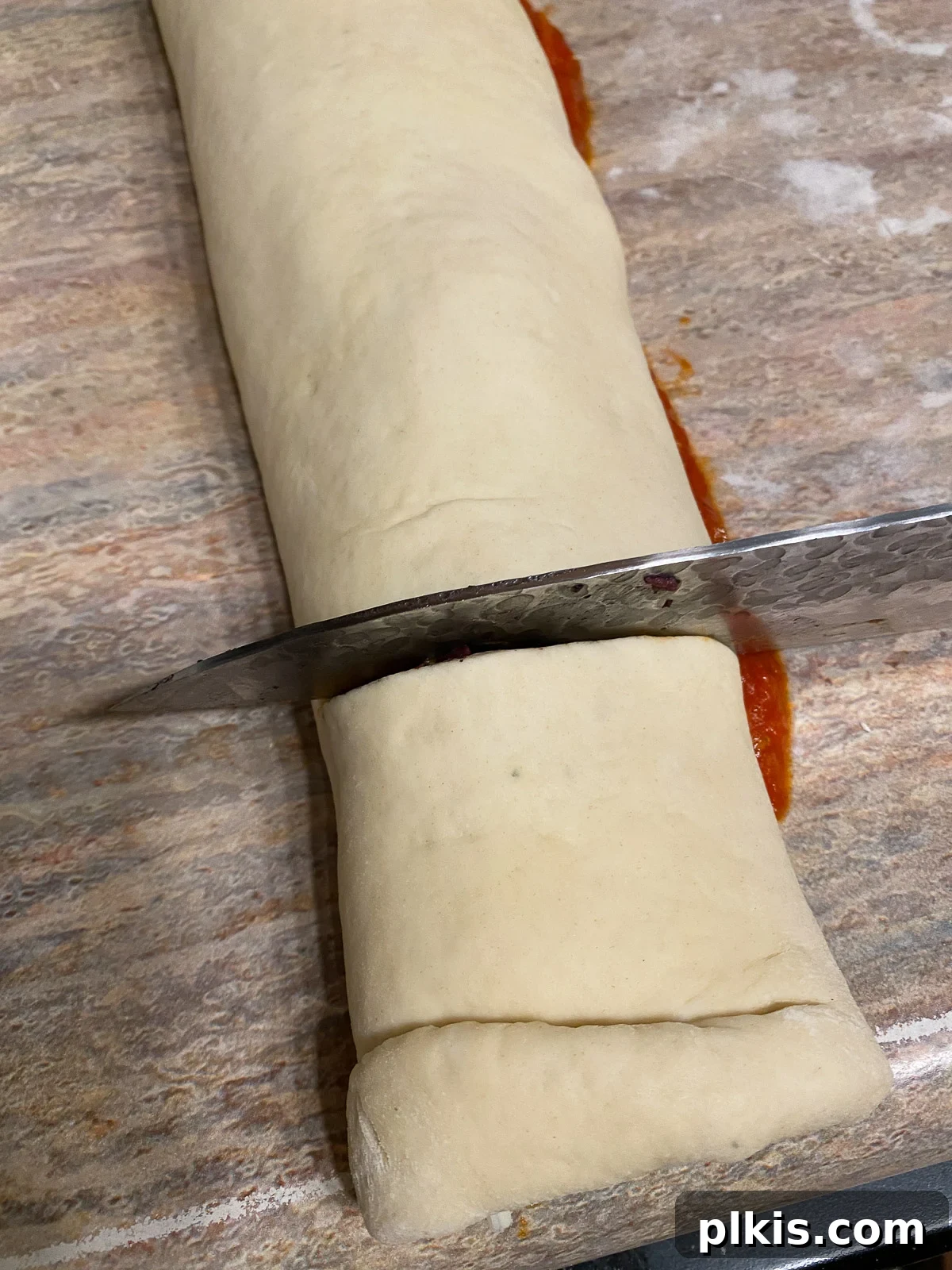 A sharp knife slicing the rolled pizza dough log into uniform 1-inch thick pizza rolls, revealing the cross-section.