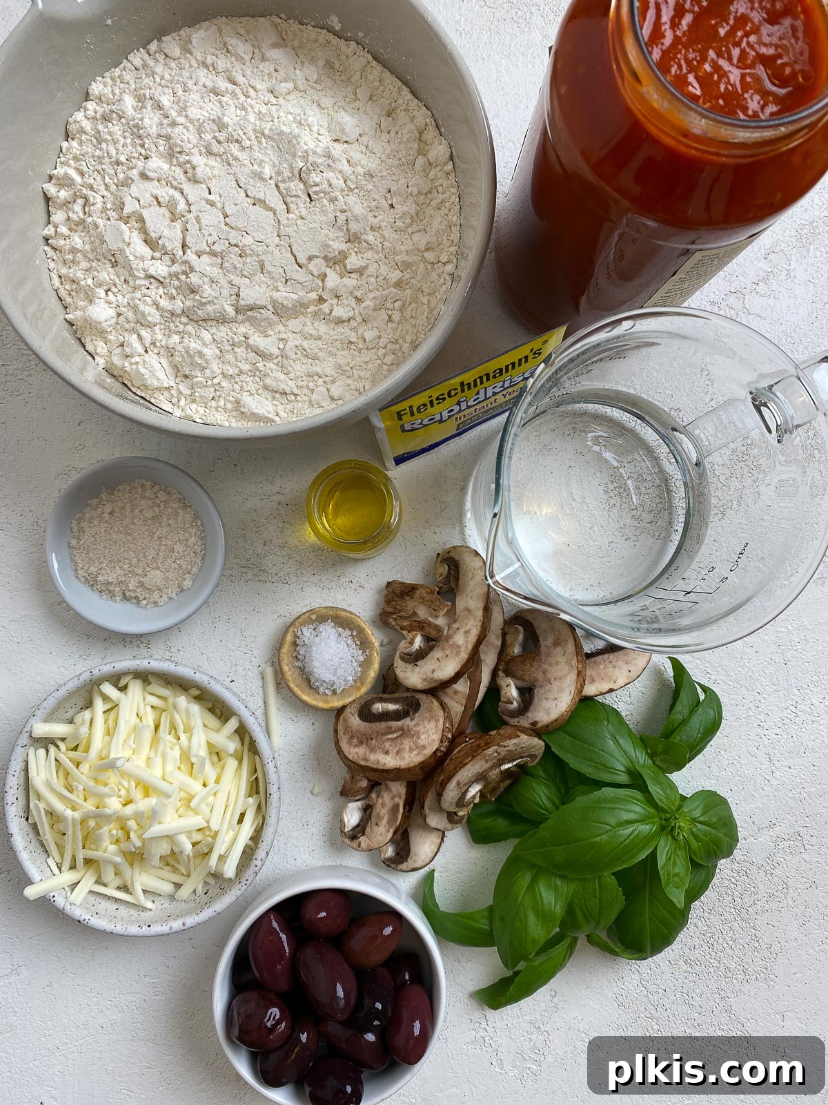 Carefully arranged ingredients for Vegan Pizza Rolls including flour, yeast, olive oil, and various colorful toppings on a clean white surface.