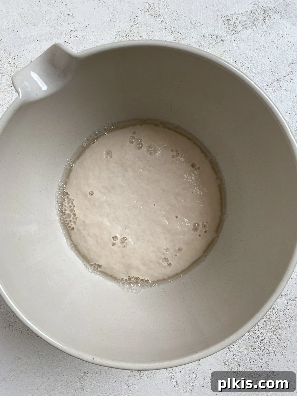 A white bowl containing warm water, active dry yeast, and sugar, showing the initial stages of yeast activation with a slight foamy layer forming.