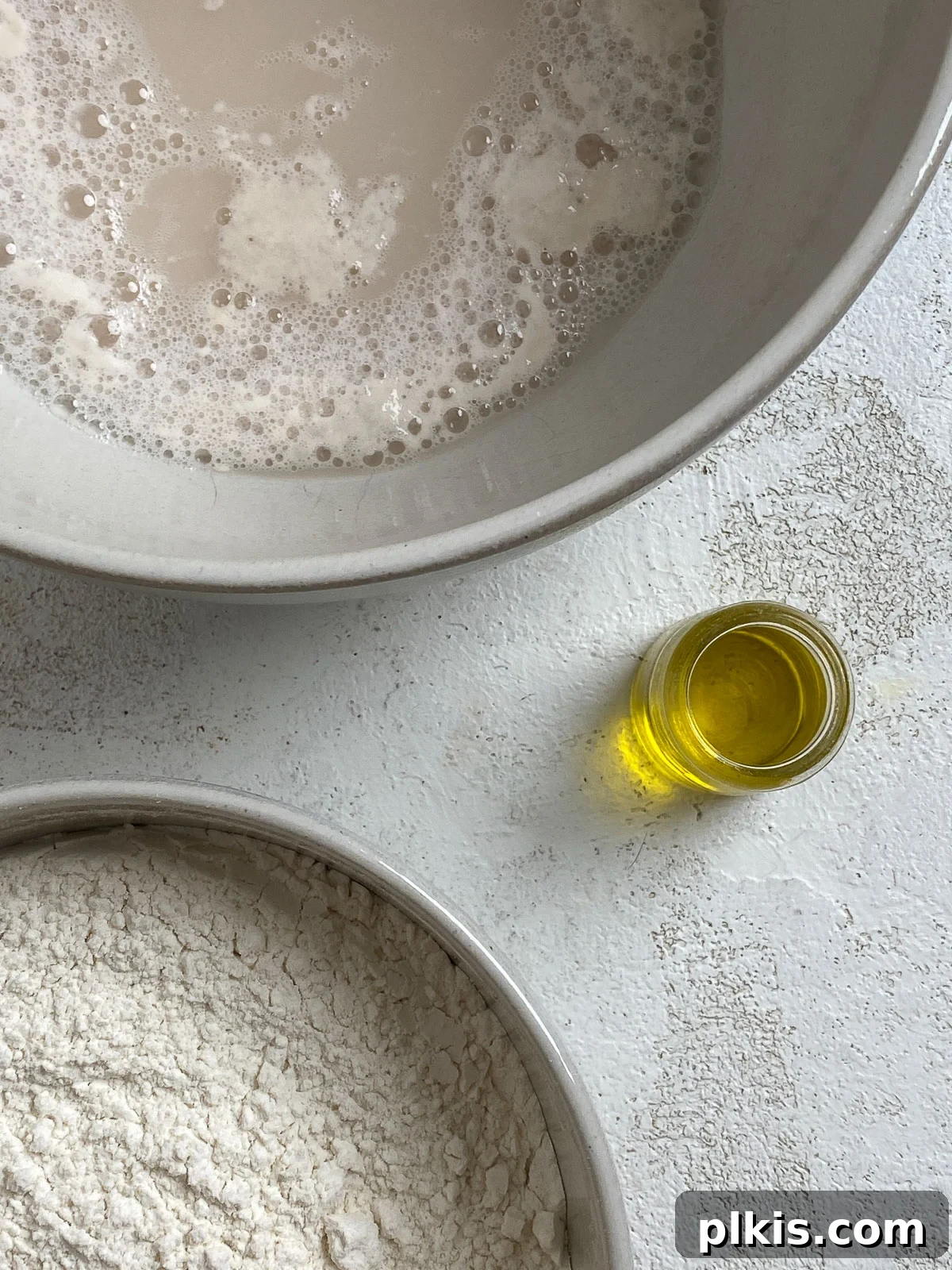 Two bowls, one with dry ingredients and one with the yeast mixture, alongside a bottle of olive oil, showcasing all components before mixing.