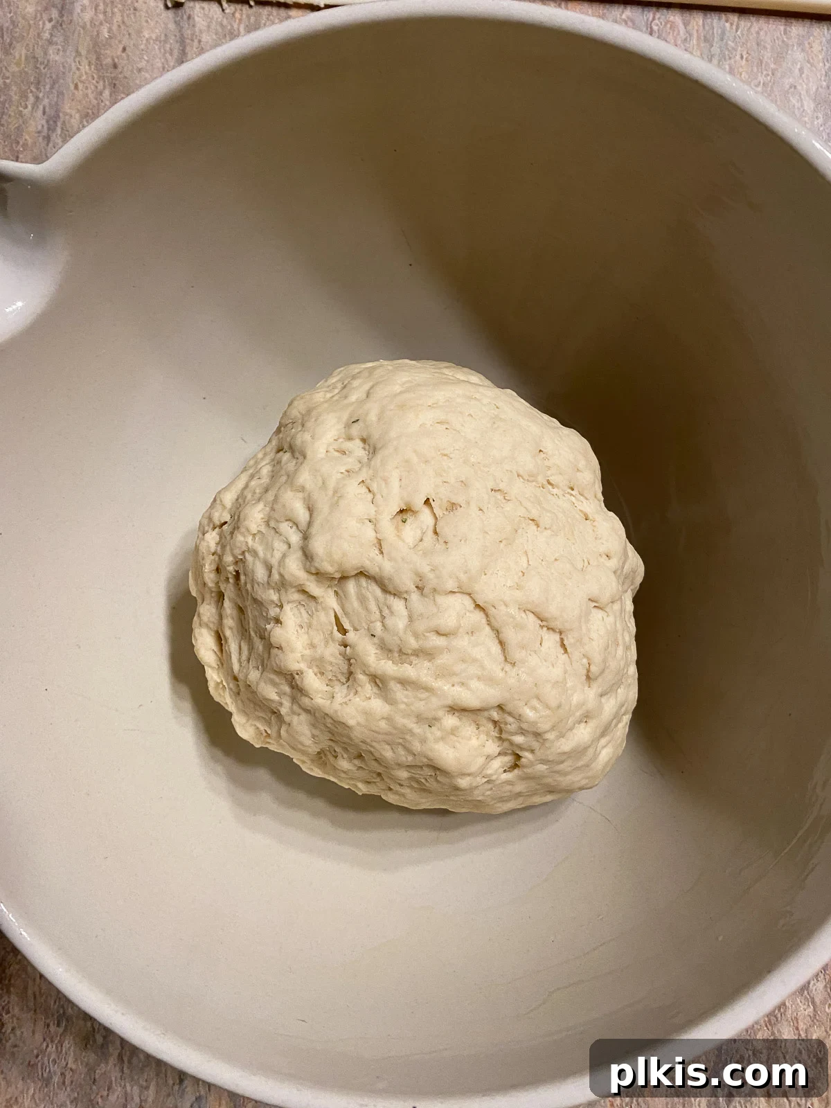 A freshly formed ball of pizza dough resting in a lightly oiled white bowl, covered, to allow it to rise.