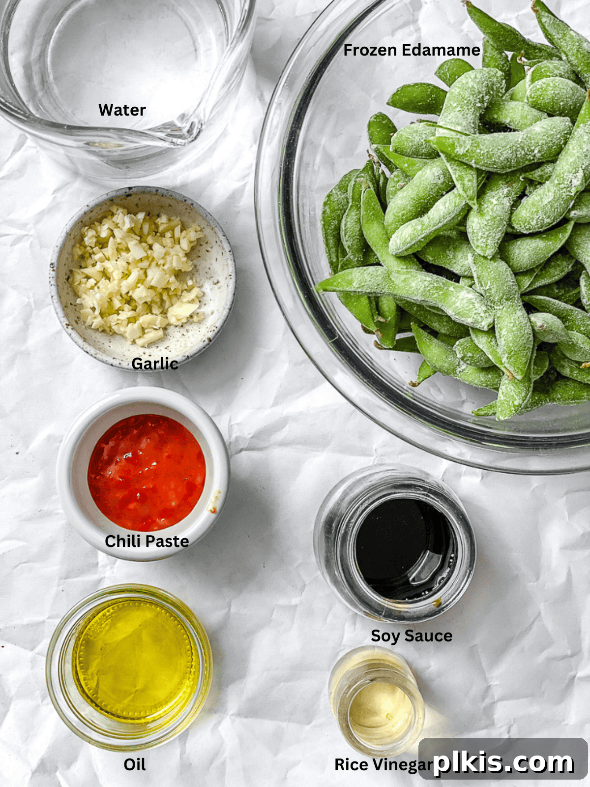 All the fresh ingredients for Quick Spicy Edamame [Chili and Garlic] are neatly laid out on a clean white surface, showcasing minced garlic, chili paste, soy sauce, rice vinegar, and fresh edamame pods.
