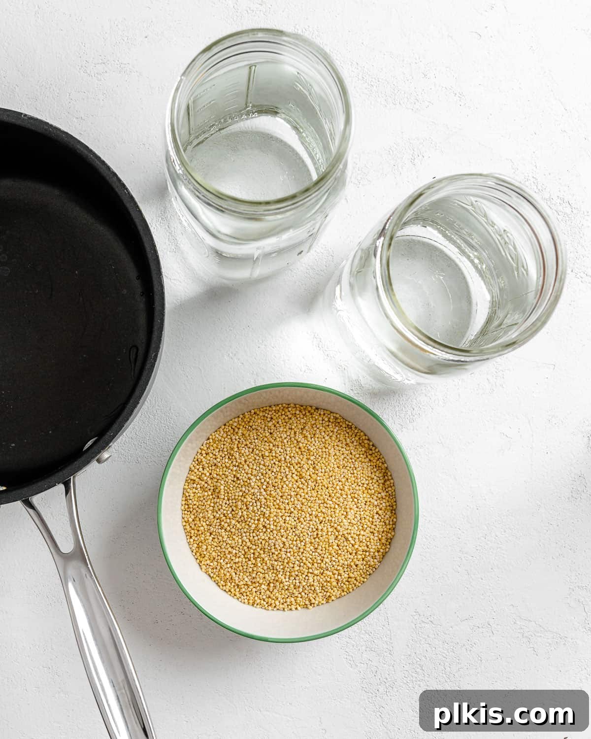 Vibrant Vegan Greek Millet Bowl 4 A cooking scene showing a black pan, two jars, and a bowl of uncooked millet, ready for preparation against a white background.