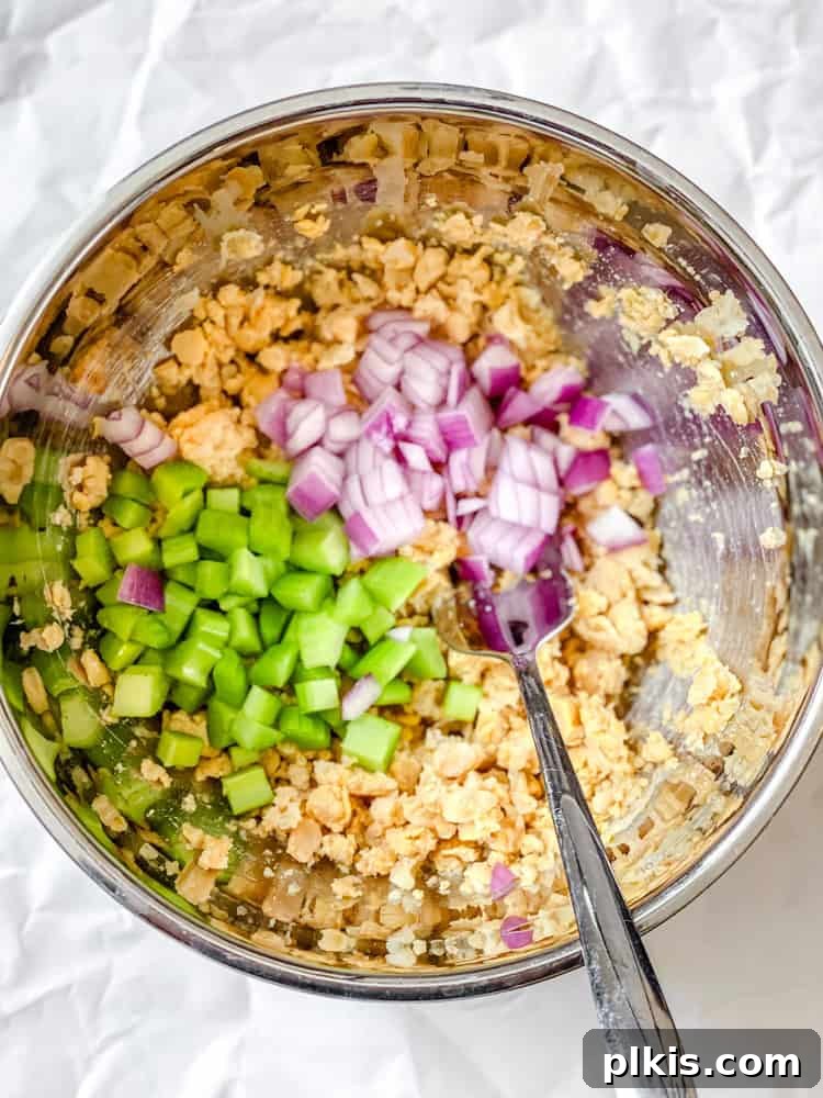 A process shot demonstrating the mixing of ingredients for Easy Curried Chickpea Salad in a bowl, capturing the initial stage of combining the mashed chickpeas with the diced vegetables before the dressing is added.