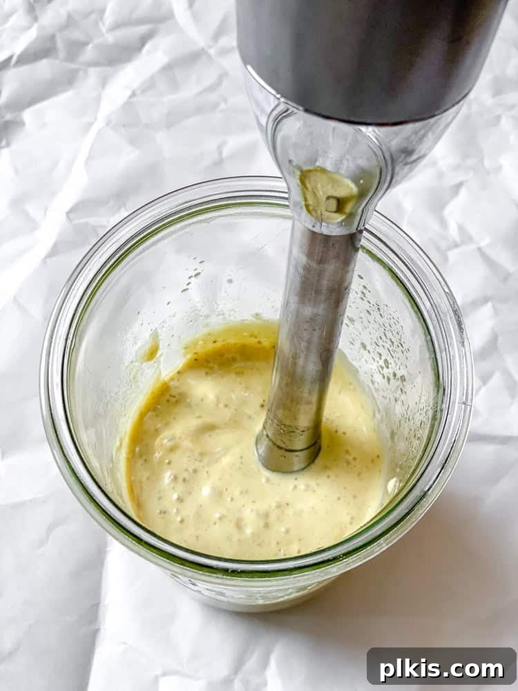A process shot illustrating the use of an immersion blender to create the creamy dressing for Easy Curried Chickpea Salad, showing the silken tofu and spices being blended to a smooth consistency in a tall jar.