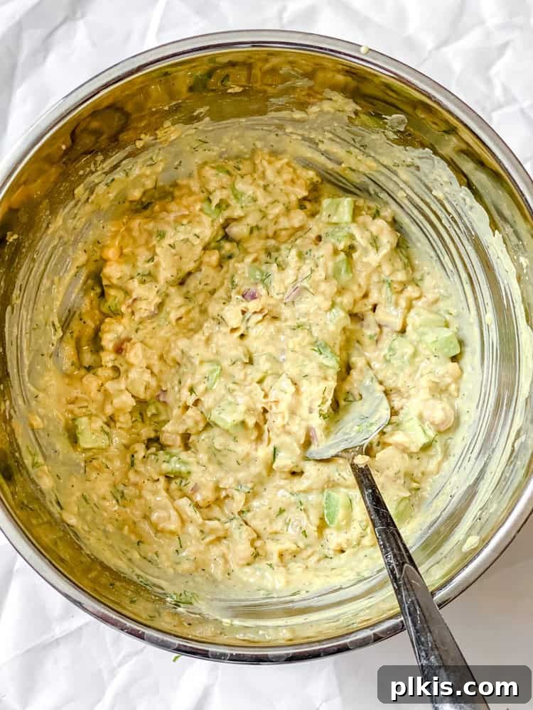 A process shot showcasing the final mixing stage of Easy Curried Chickpea Salad, with the creamy dressing and fresh dill being stirred into the mashed chickpea and vegetable mixture in a bowl, ensuring all ingredients are thoroughly combined.