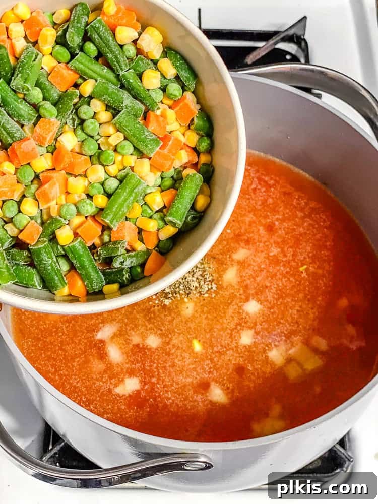 Process shot illustrating the addition of mixed vegetables and diced tomatoes to the simmering broth in a large pot.