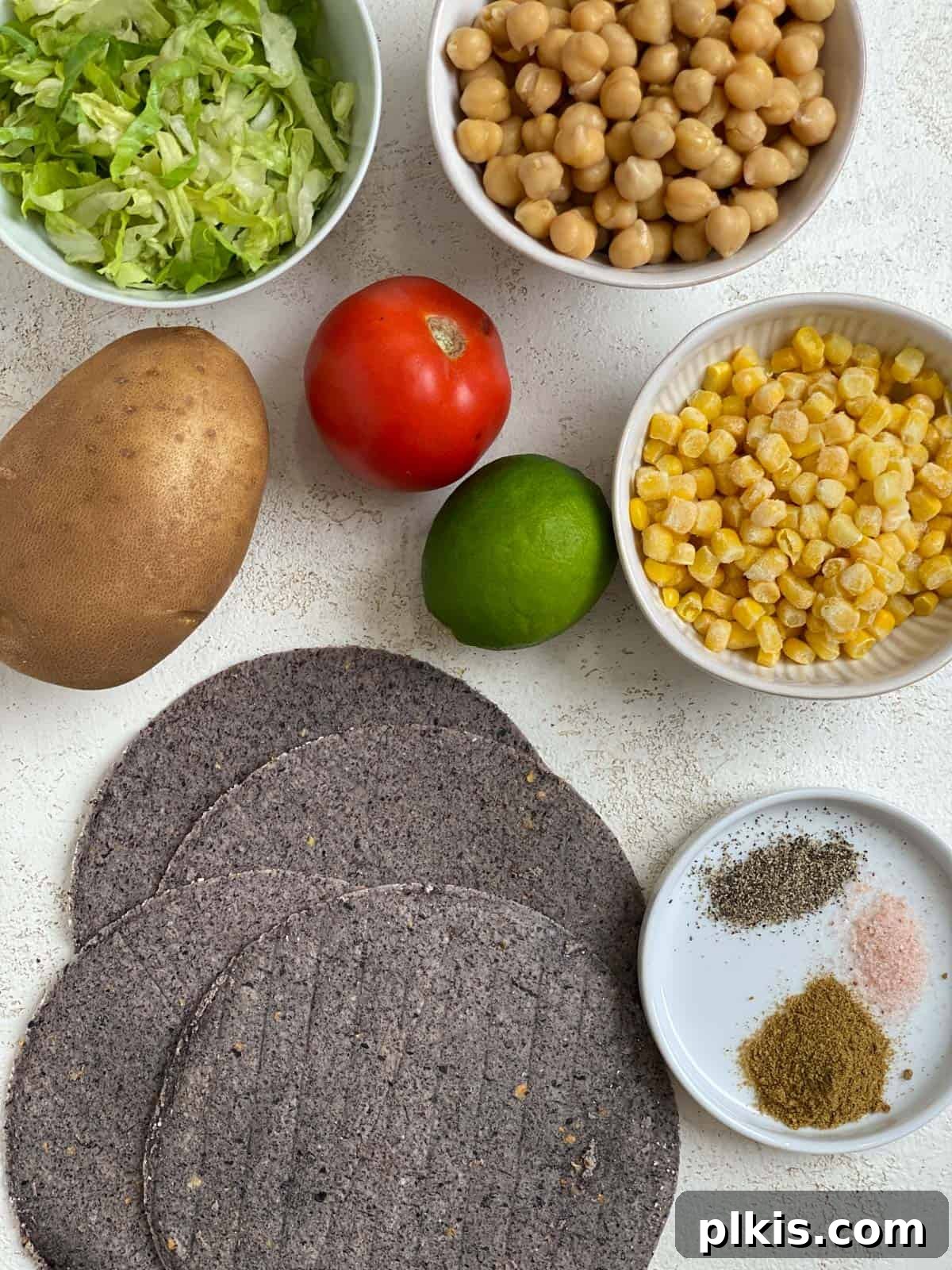 Golden Potato and Chickpea Tacos 4 Close-up view of fresh ingredients for potato and chickpea tacos: bright red tomatoes, crisp lettuce, yellow corn kernels, a whole lemon, and a bottle of hot sauce, against a light backdrop.