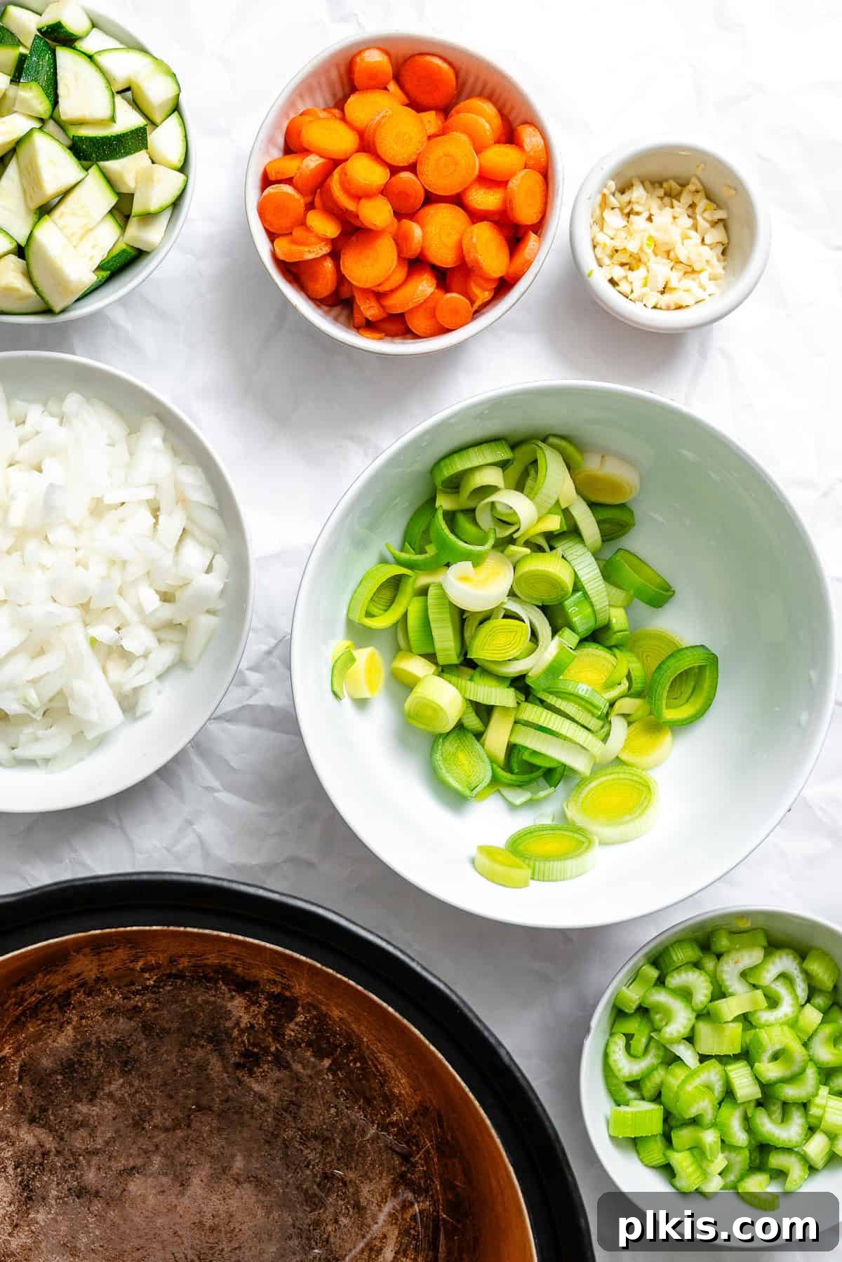 Hearty Vegan Potato and Vegetable Soup 4 Prepped vegetables for soup, including diced onion, sliced leeks, carrots, zucchini, and minced garlic, arranged in separate bowls on a white surface.