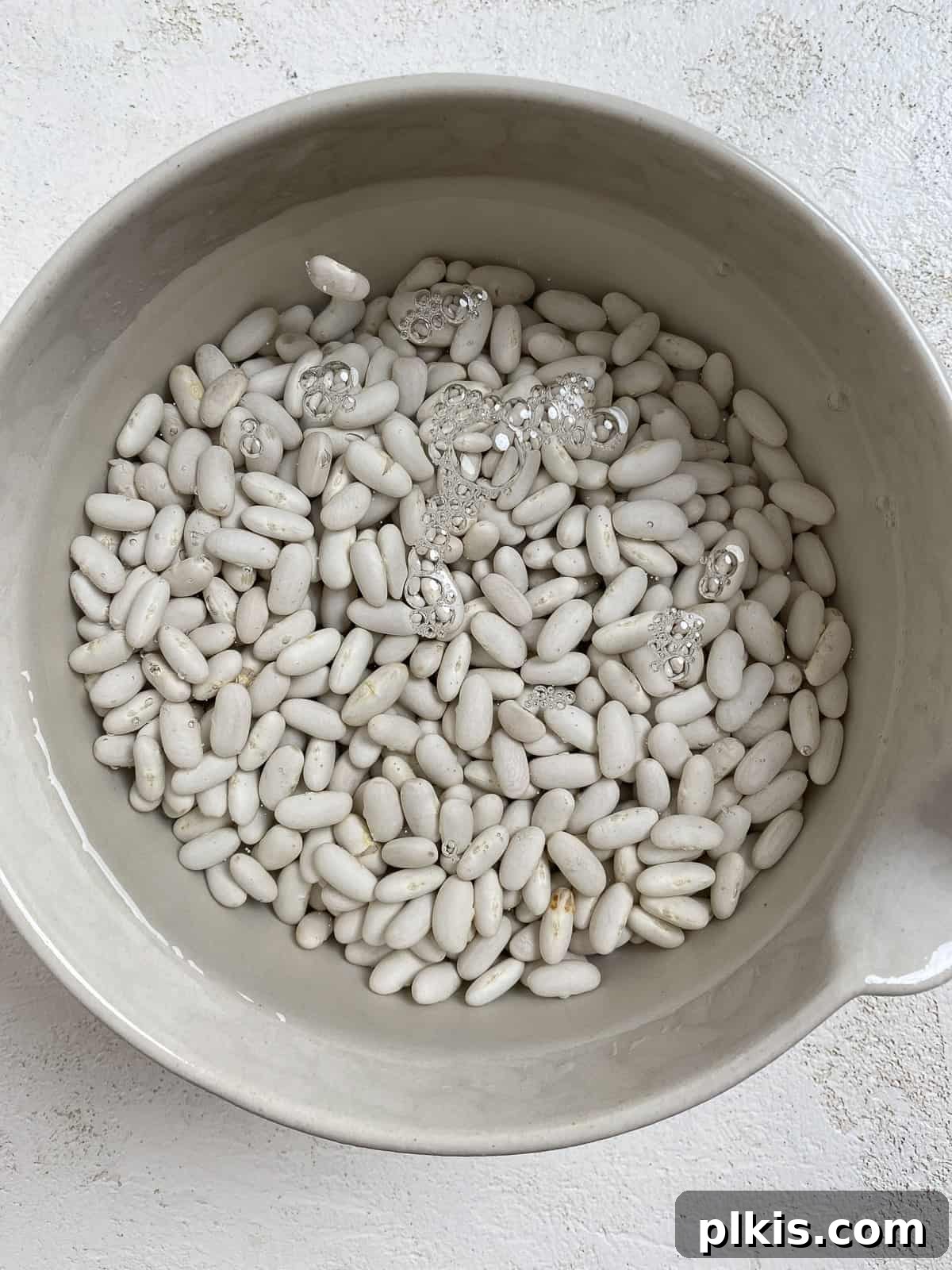 Homestyle Vegan Slow Cooker Baked Beans 4 Process shot of dried white beans soaking in a bowl of water on a white surface
