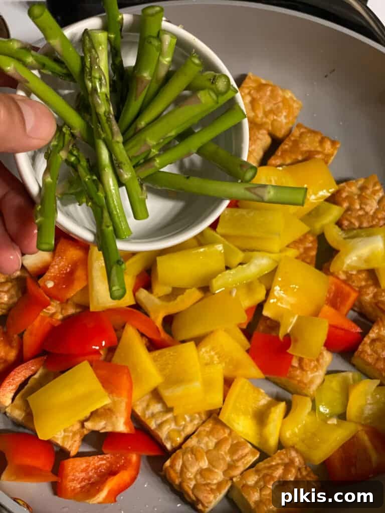 Process shot of adding asparagus spears to the pan with tempeh and bell peppers.