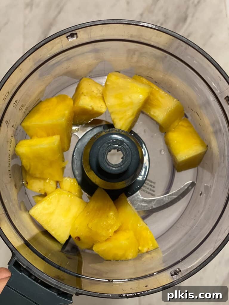 Process shot of fresh pineapple chunks being added to a food processor bowl.