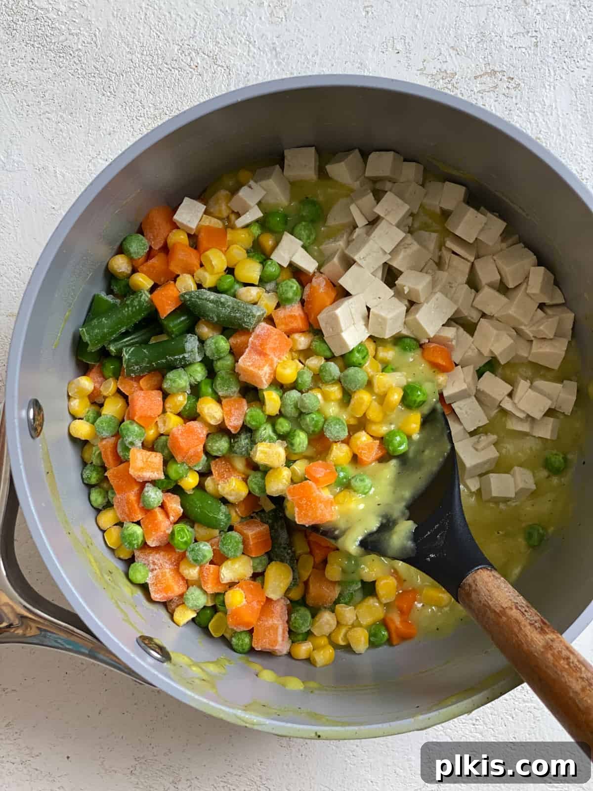Close-up process shot of vibrant mixed vegetables and tofu being stirred into creamy vegan gravy in a mixing bowl, creating the pot pie filling.