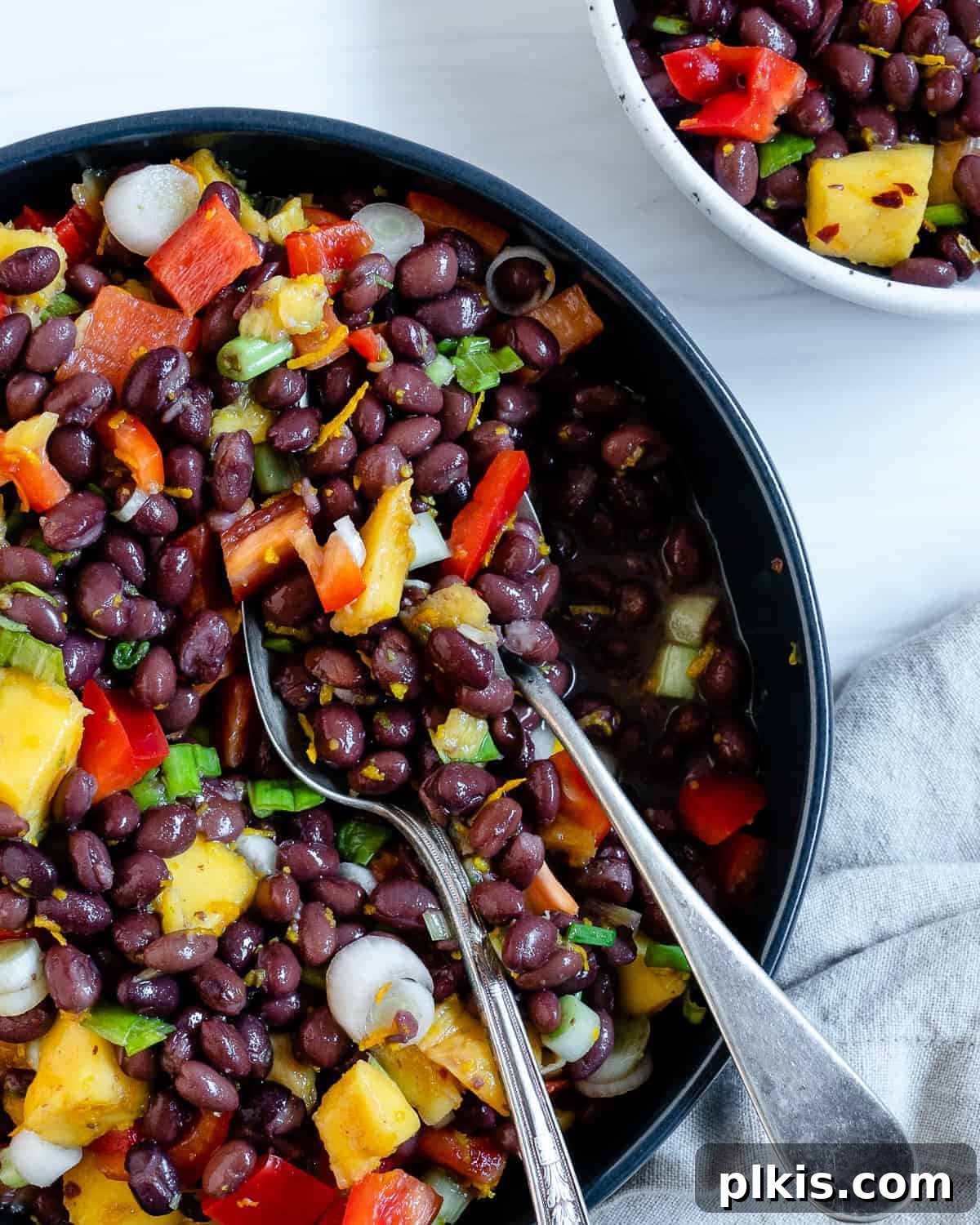 A vibrant mango black bean salad in a dark bowl, ready to be served. Utensils rest inside the bowl against a clean white background.