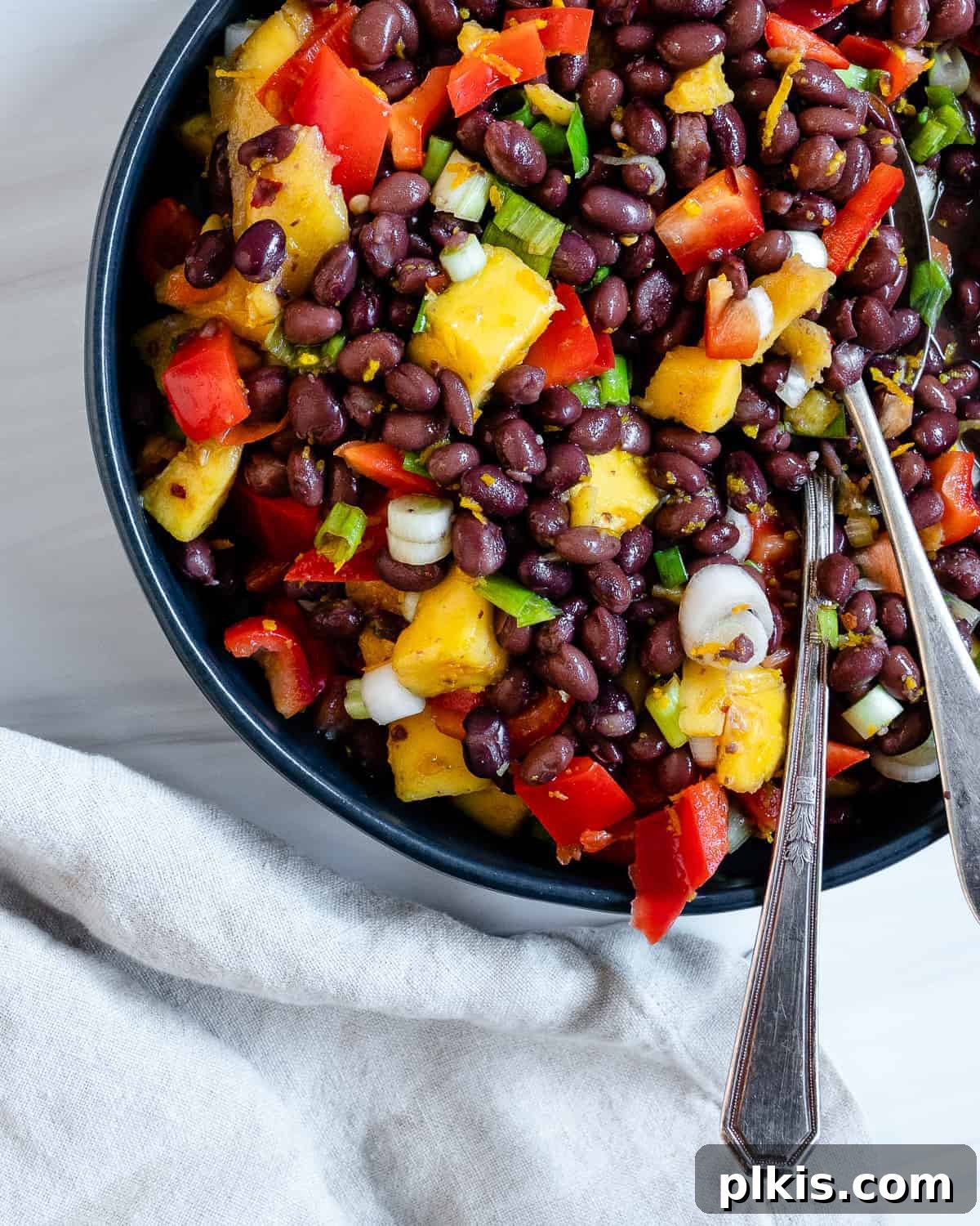 Another view of the completed mango black bean salad in a dark bowl, with serving utensils, against a clean white background.