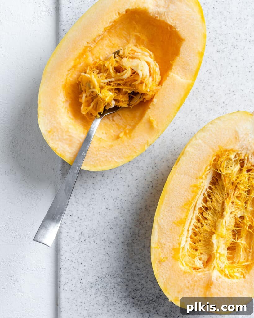 The Secret to Delicious Baked Spaghetti Squash 4 Process shot showing a spoon scooping out the seeds and pulp from inside a halved spaghetti squash.