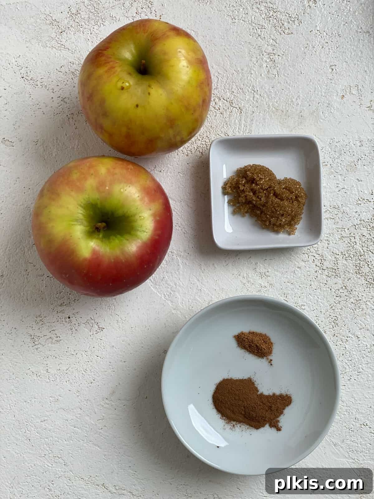 Crispy Apple Slices 3 A top-down view of fresh ingredients for baked apple chips on a white surface, including crisp apples, a bowl of cinnamon sugar, and a mandoline slicer.