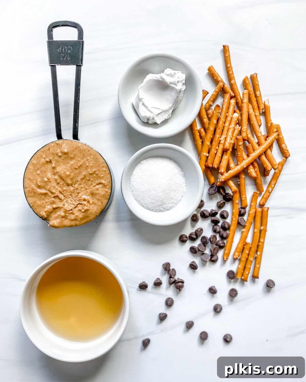 Frightfully Delicious Peanut Butter Spiders 3 Ingredients for peanut butter spiders laid out on a white surface, including peanut butter, sugar, flour, pretzels, and chocolate chips.