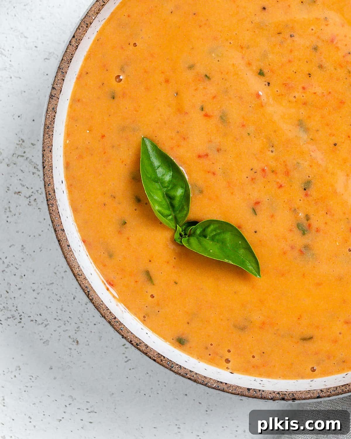 Velvety Roasted Red Pepper Soup 9 A beautifully presented bowl of creamy roasted red pepper soup, garnished with fresh basil leaves, on a light, minimalist surface.